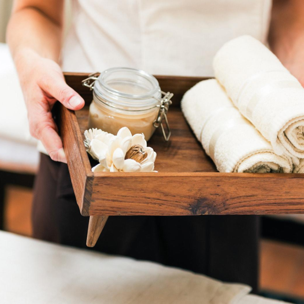 Spa therapist holding a wooden tray with white towels, a jar, and a white flower